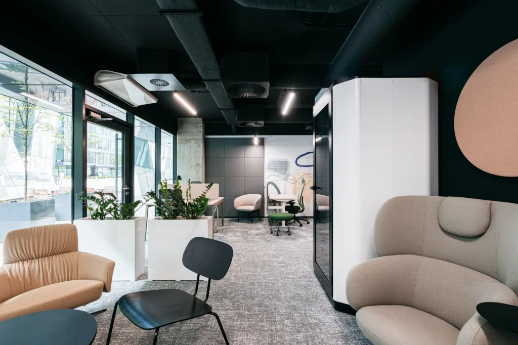 Okrągły panel akustyczny z filcu na ścianie oraz wolnostojąca kabina dźwiękoszczelna w nowoczesnym biurze z ergonomicznymi meblami i strefą relaksu.