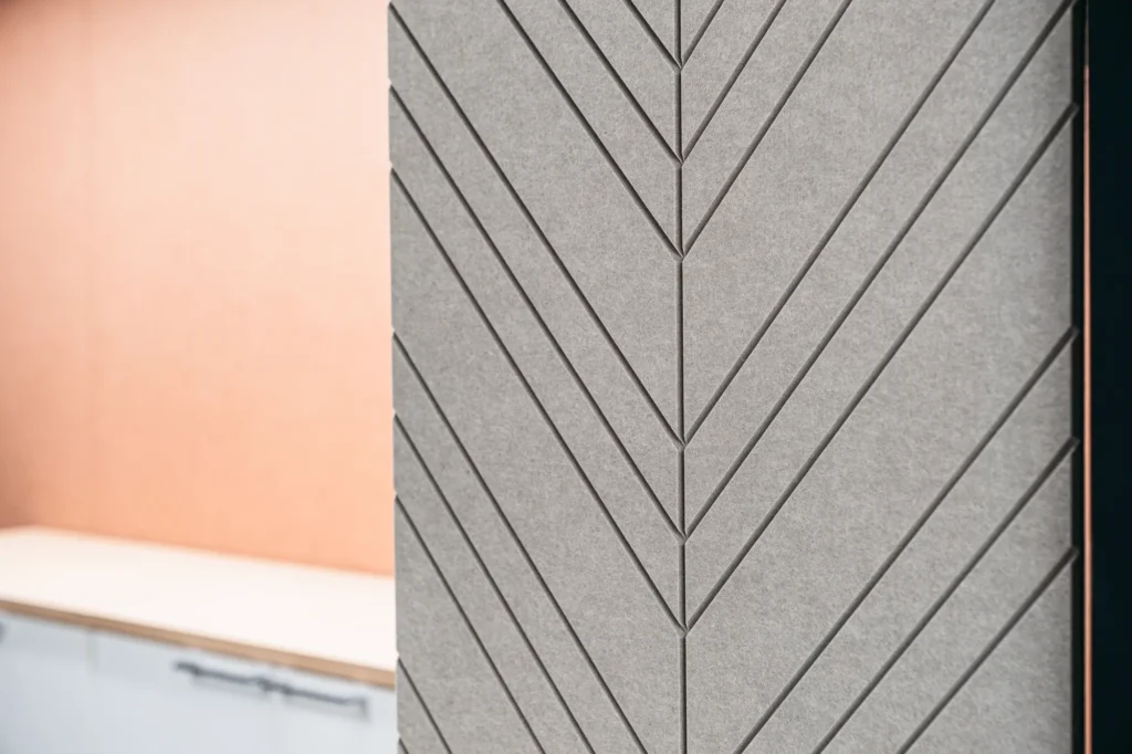 A close-up of a gray acoustic felt panel with a milled herringbone pattern, mounted on a section of a wall or pillar, with a blurred orange office background.
