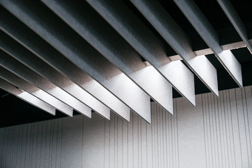 A suspended acoustic ceiling made of parallel, vertically suspended felt panels in shades of gray, illuminated by linear LED lighting.
