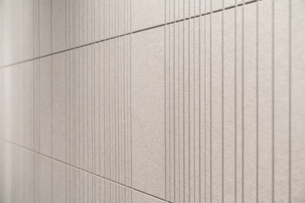 A close-up shot of light gray acoustic felt wall panels with precise vertical slits forming a rhythmic pattern of lines.